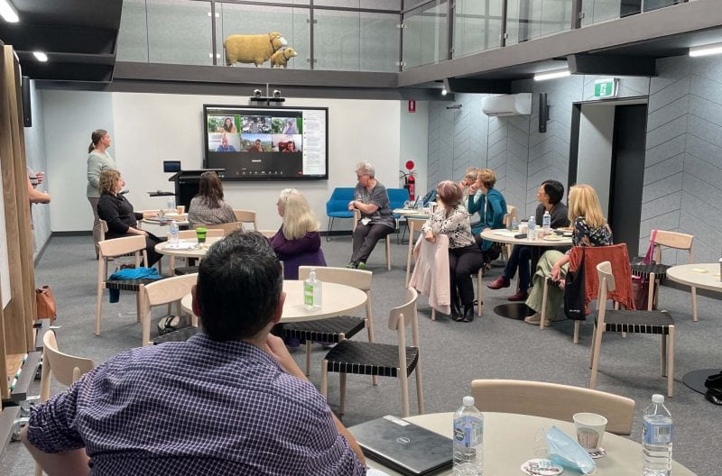 A group of UNE staff gathered in NOVA. They are seated around tables and all are looking to the front of the room where a number of participants are shown on screen via Zoom. A model sheep can be seen on the walkway above the room.
