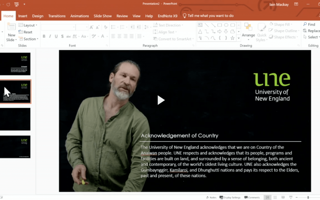 A screenshot of a Powerpoint file overlaid over Iain. Iain is demonstrating how the overlay works. The slide displays the UNE logo and an Acknowledgement of Country.