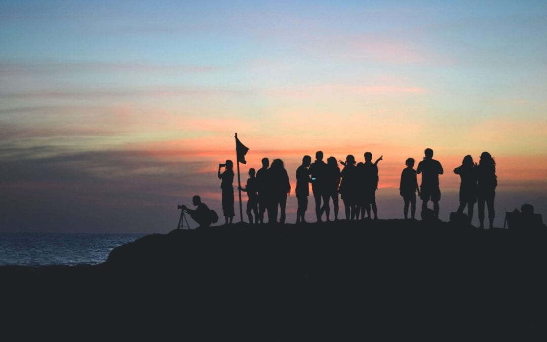 A group of people with a flag at the summit of a mountain, silhouetted against a sunset.