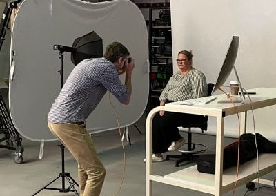 Dee Dell sits in a photography studio posing for a profile photo while Matt Cawood points the camera in her direction.