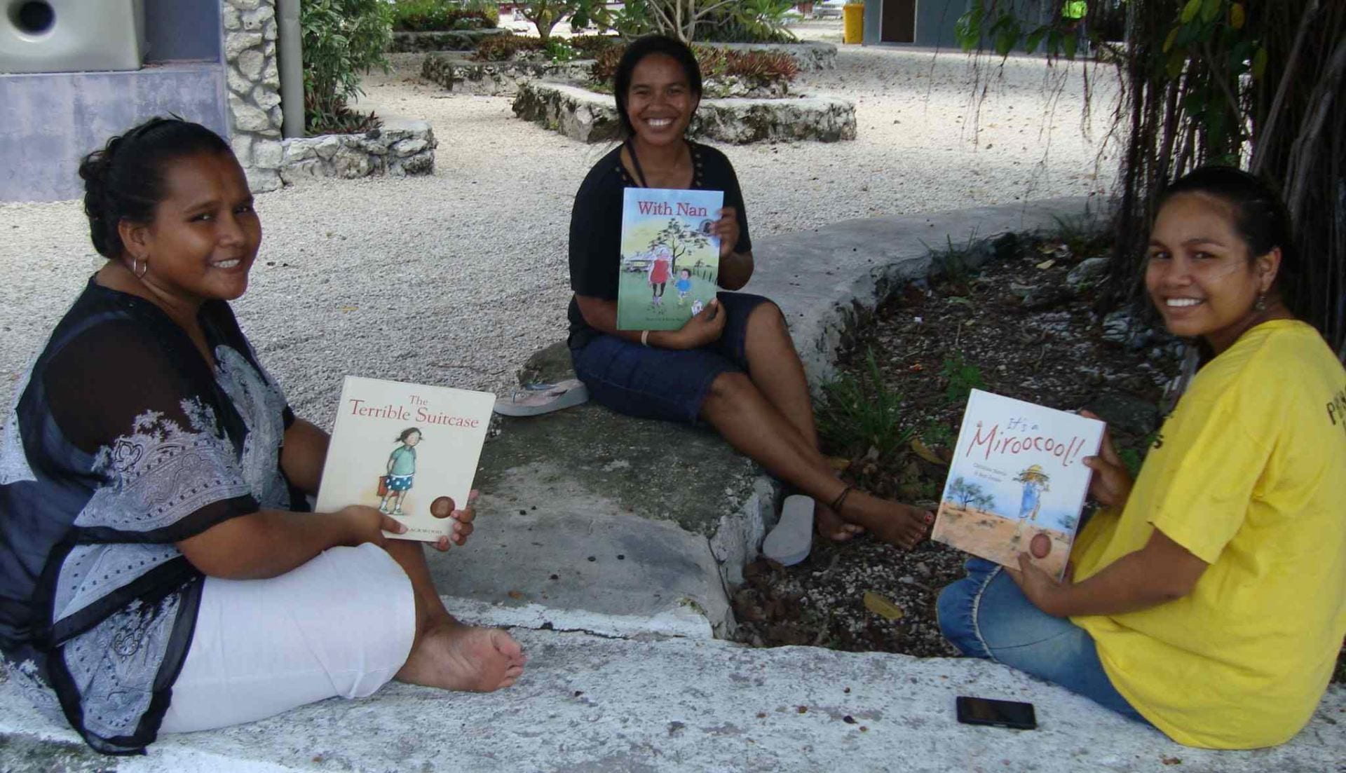 Three smiling Nauruan university students sit outdoors each holding a children's book in English
