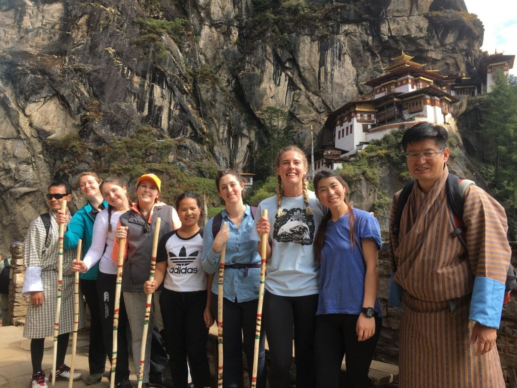 Students and their guides in traditional Bhutanese dress in front of a temple in a cliff side.