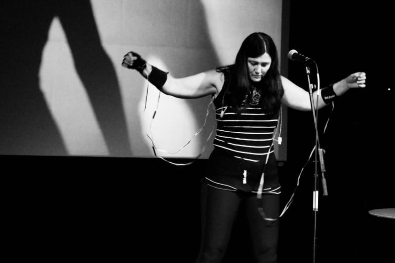 Black-and-white image of Donna Hewitt on stage hooked up to electronic wearables with arms outstretched