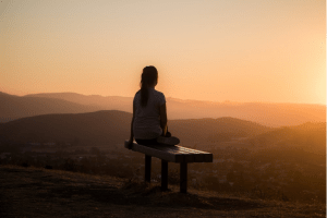 Woman sitting on bench seat cross-legged looking at horizon at sunset