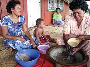 Islander women preparing food in an outdoor setting, with a child sitting nearby.