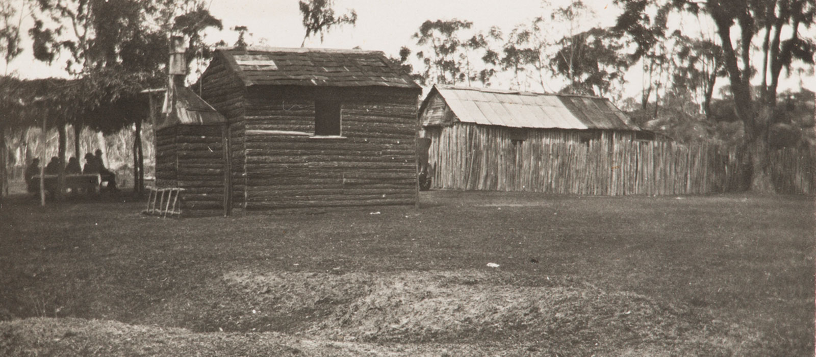 Historic black and white photo of log cabins and a group of people sitting under a shelter in the Australian bush.