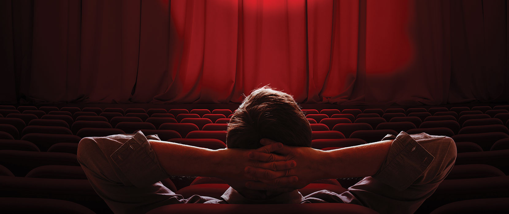 Theatre with red curtain pulled over a stage, with a sole man sitting in the audience with his hands behind his head, as if ready to enjoy a show.