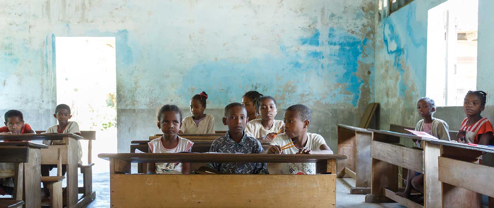 Malagasy school children sitting on desks in a basic room with rough plaster on the walls.