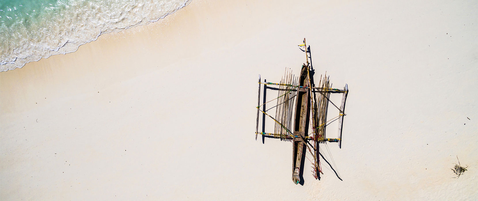 Aerial image of outrigger canoe on a sandy beach with turquois sea.