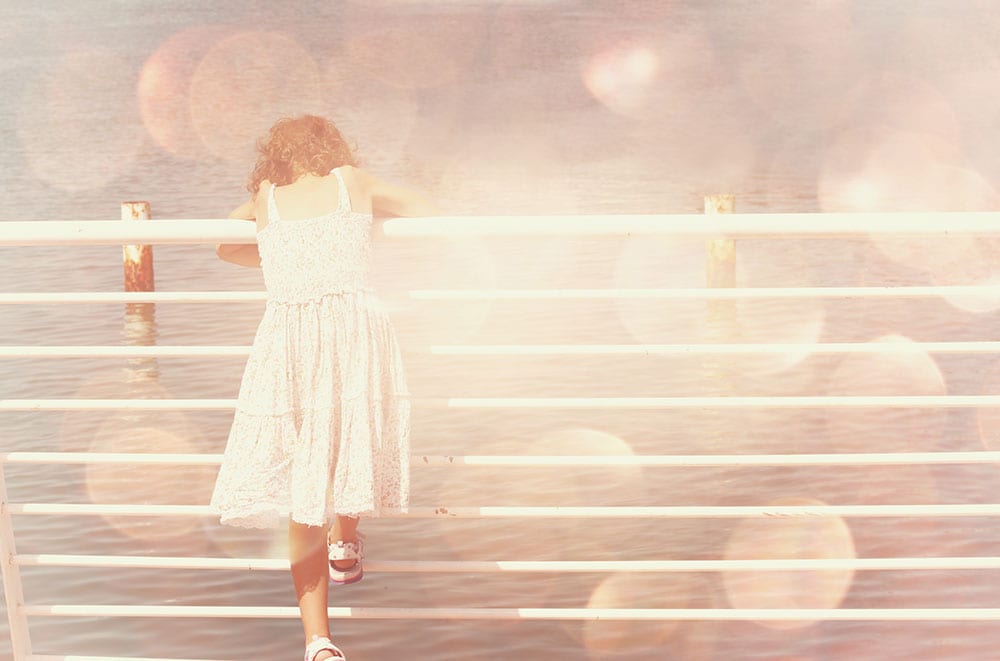 Young girl leaning over a railing on a pier, looking at the sea. Pink and peachy hues.